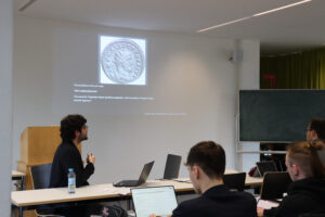 Eine Person hält einen Vortrag in einem Klassenzimmer und zeigt eine Folie mit einer antiken Münze und einem Text. Mehrere Personen sitzen an Tischen mit Laptops und hören aufmerksam zu.