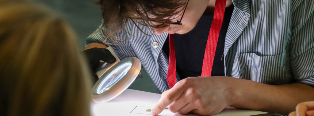 Eine Person, die eine Brille trägt, untersucht mit einer Lupe ein kleines Objekt auf einem Blatt Papier, wobei sie mit einem Finger auf das Papier zeigt und ein rotes Schlüsselband um den Hals trägt. Eine Person, die eine Brille trägt, untersucht mit einer Lupe ein kleines Objekt auf einem Blatt Papier, wobei sie mit einem Finger auf das Papier zeigt und ein rotes Schlüsselband um den Hals trägt.